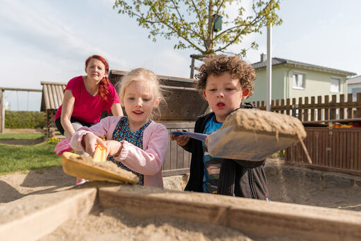 Zwei Kinder spielen gemeinsam im Sand unter Aufsicht einer Betreuerin